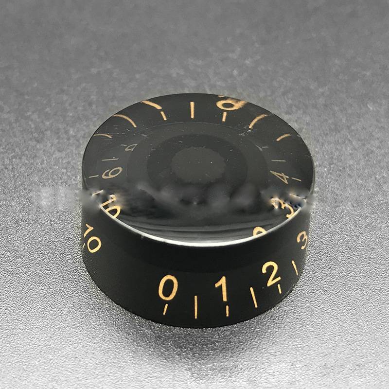 A close-up image of a black electric guitar bass volume knob with gold numerals and white lettering.