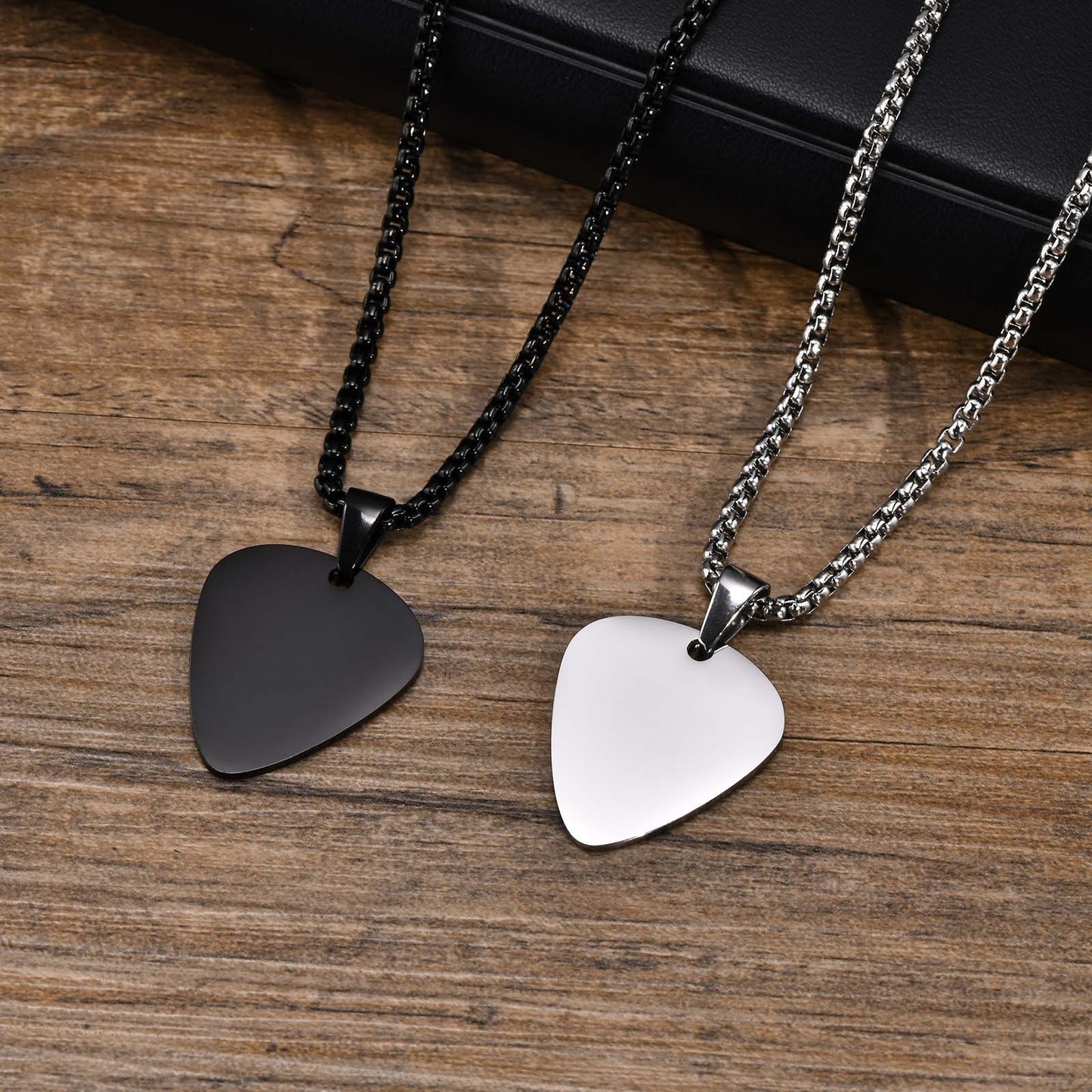 Two guitar pick pendants, one black and one silver, attached to chains laid out on a wooden surface.
