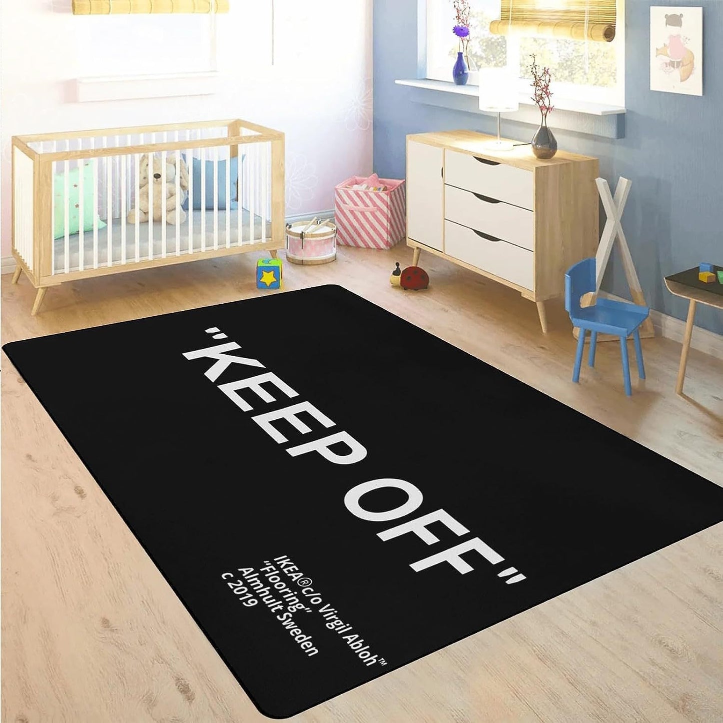 A large black carpet with white text 'KEEP OFF' printed on it, placed on a wooden floor in a room with a crib, chair, and a small table. The carpet is intended for use in a music room.