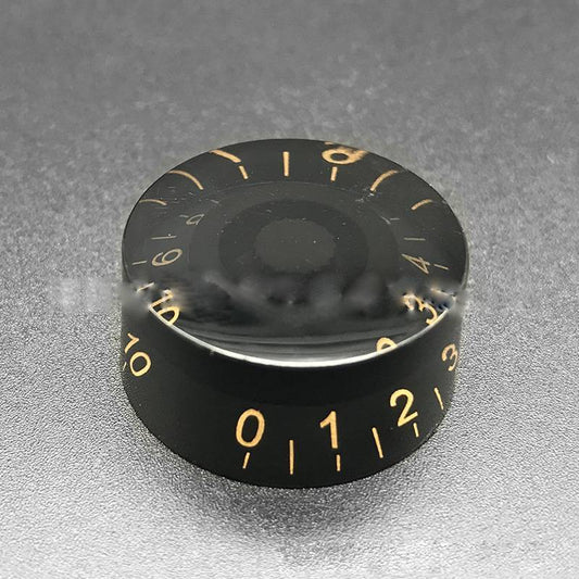 A close-up image of a black electric guitar bass volume knob with gold numerals and white lettering.